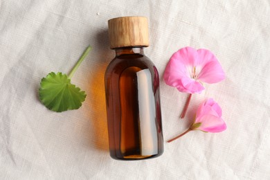 Photo of Bottle of geranium essential oil and beautiful flowers on white fabric, flat lay