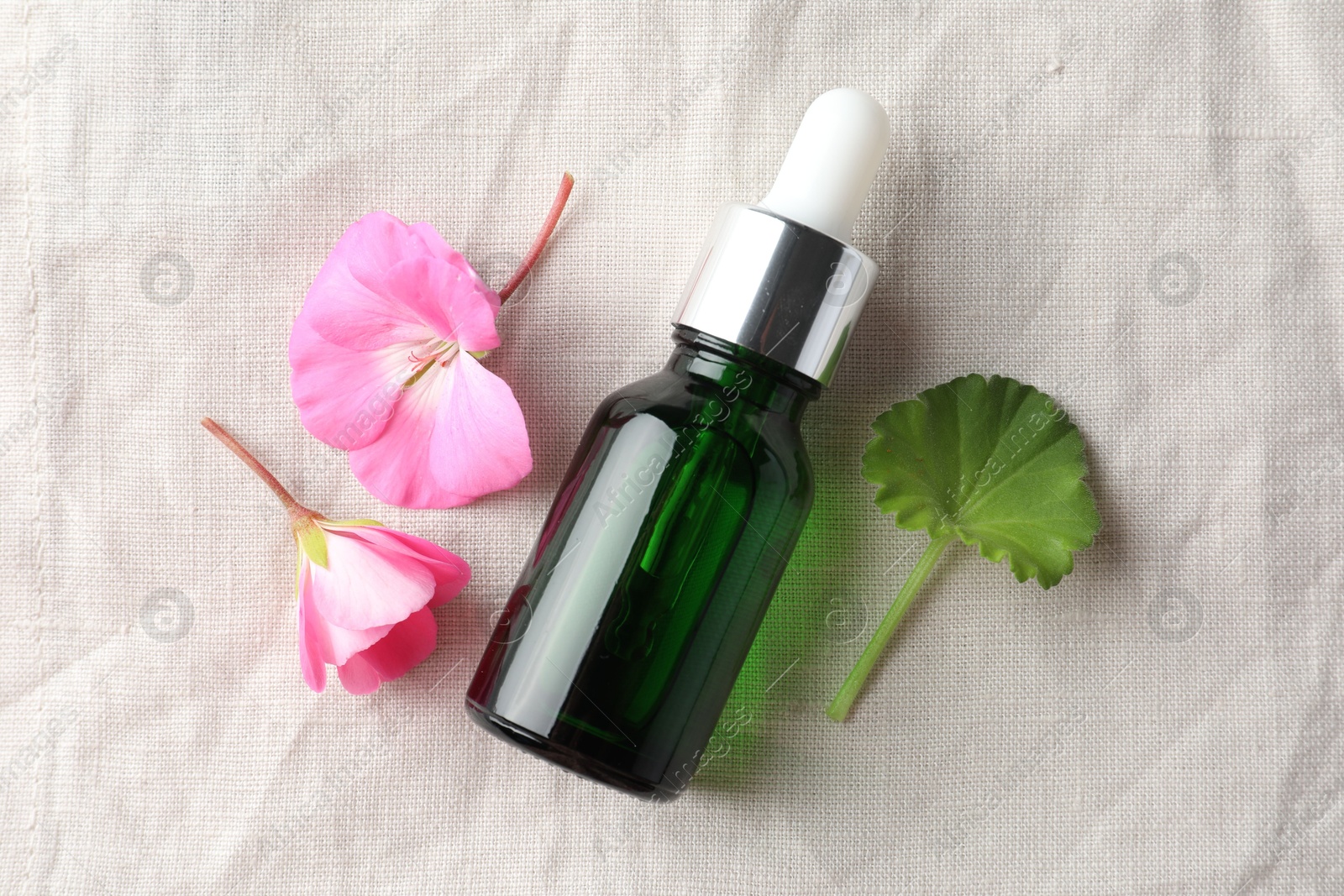 Photo of Bottle of geranium essential oil and beautiful flowers on white fabric, flat lay