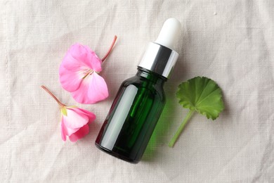 Photo of Bottle of geranium essential oil and beautiful flowers on white fabric, flat lay