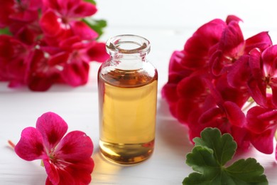 Photo of Bottle of geranium essential oil and beautiful flowers on white wooden table, closeup