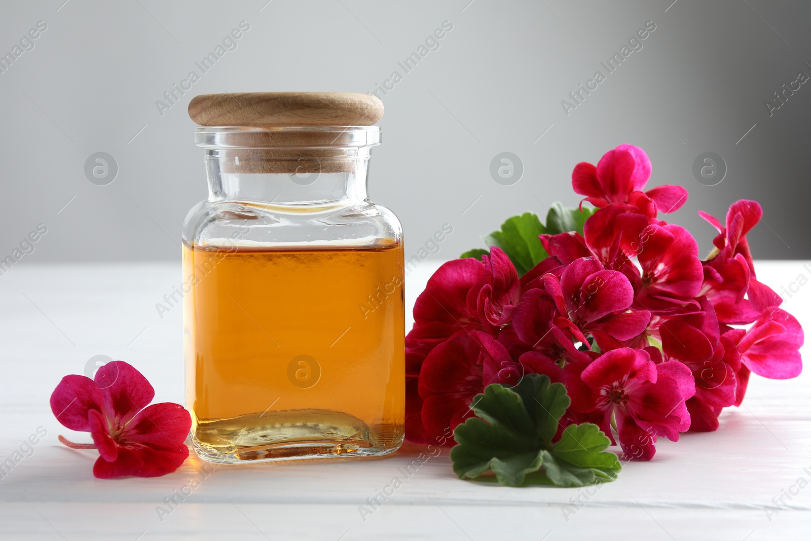 Photo of Bottle of geranium essential oil and beautiful flowers on white wooden table, closeup