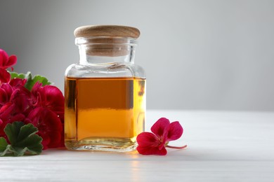 Photo of Bottle of geranium essential oil and beautiful flowers on white wooden table, closeup. Space for text