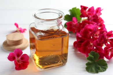 Photo of Bottle of geranium essential oil and beautiful flowers on white wooden table, closeup