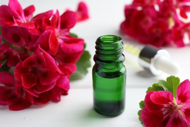 Photo of Bottle of geranium essential oil and beautiful flowers on white wooden table, closeup