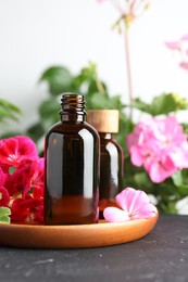 Photo of Bottles of geranium essential oil and beautiful flowers on black table, closeup