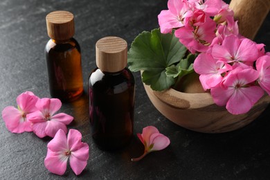 Photo of Bottles of geranium essential oil, beautiful flowers and mortar with pestle on black table, closeup