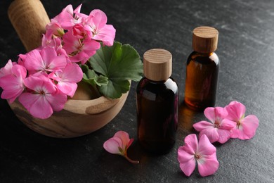 Photo of Bottles of geranium essential oil, beautiful flowers and mortar with pestle on black table