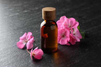 Photo of Bottle of geranium essential oil and beautiful flowers on black table, closeup