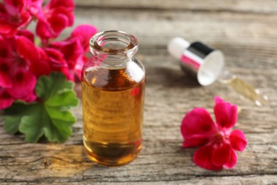 Photo of Bottle of geranium essential oil, pipette and beautiful flowers on wooden table, closeup