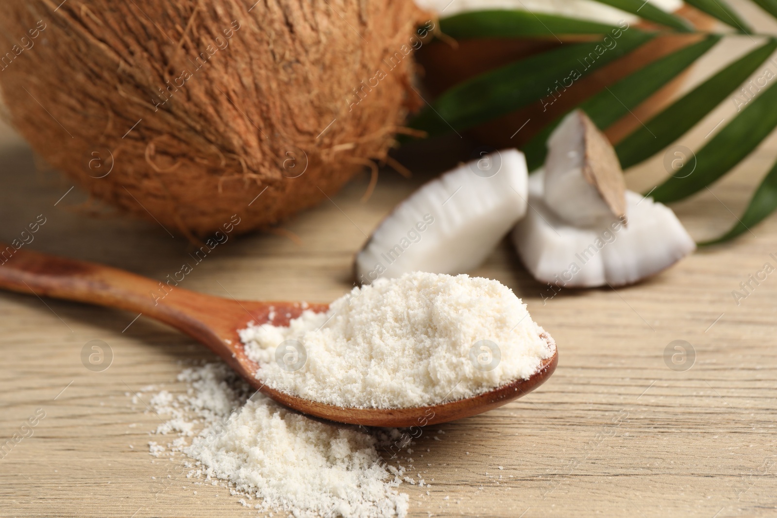 Photo of Fresh coconut flour in spoon, nuts and palm leaf on wooden table, closeup