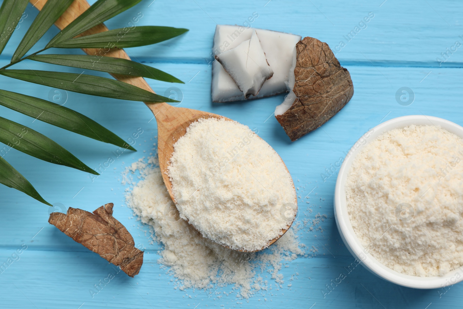 Photo of Fresh coconut flour in spoon, nut and palm leaf on light blue wooden table, flat lay