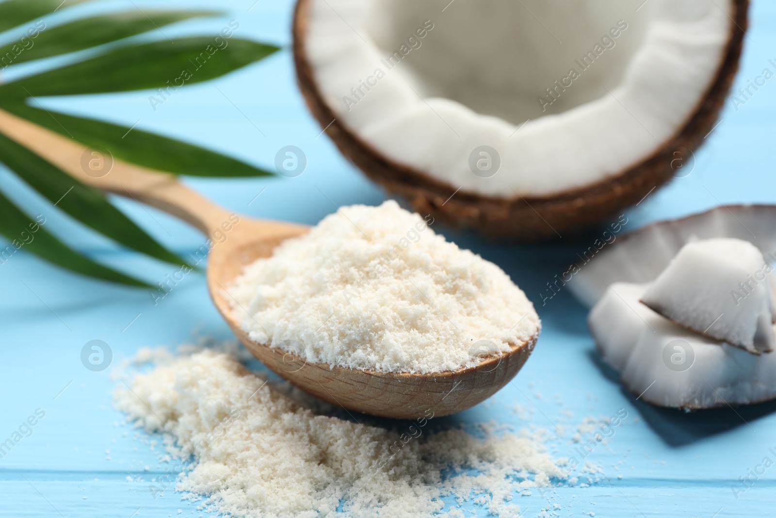 Photo of Fresh coconut flour in spoon, nut and palm leaf on light blue wooden table, closeup