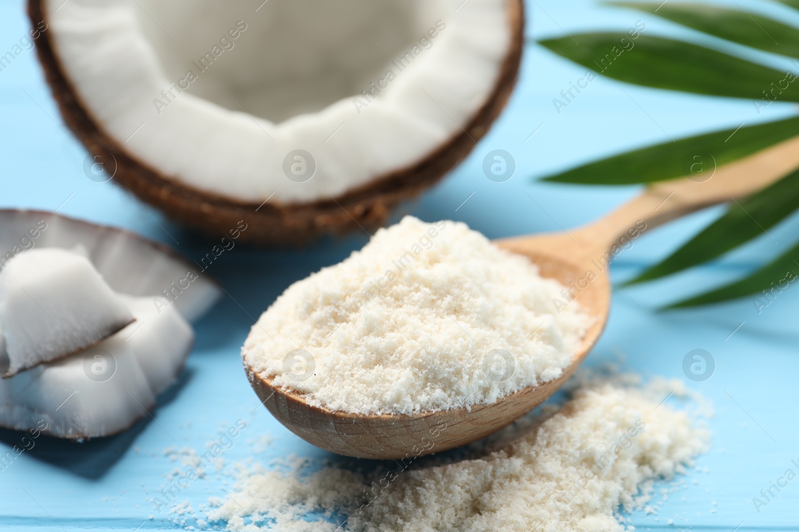 Photo of Fresh coconut flour in spoon, nut and palm leaf on light blue wooden table, closeup