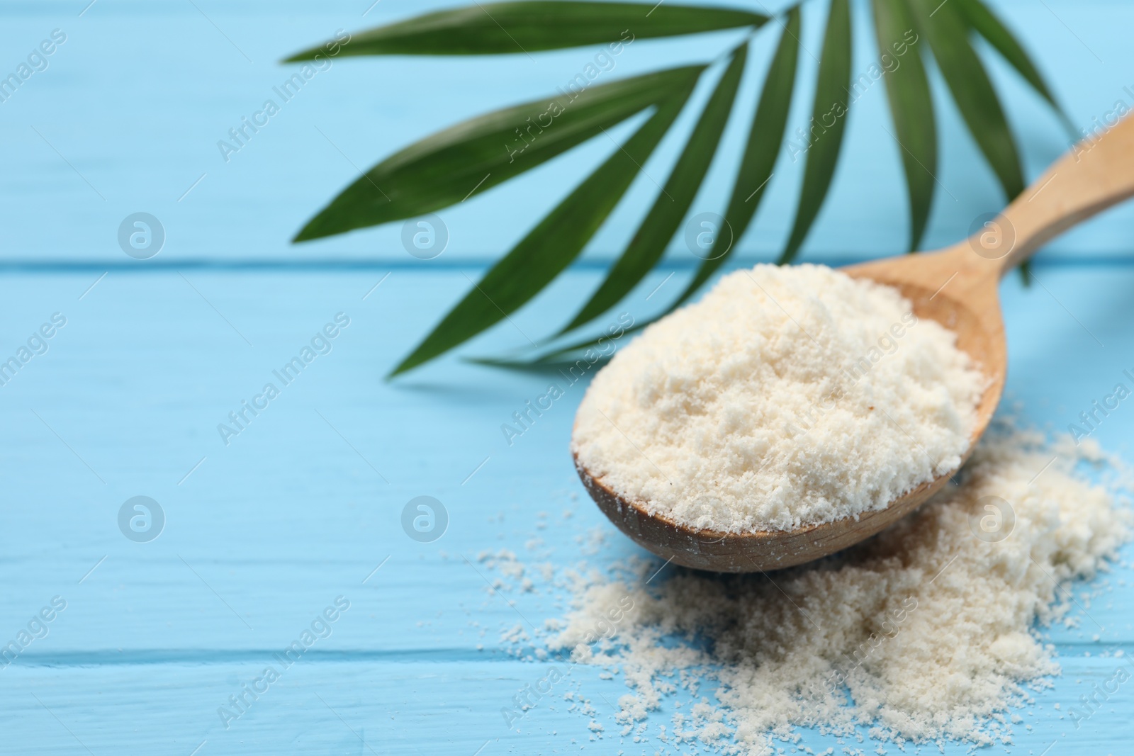 Photo of Fresh coconut flour in spoon and palm leaf on light blue wooden table, closeup. Space for text