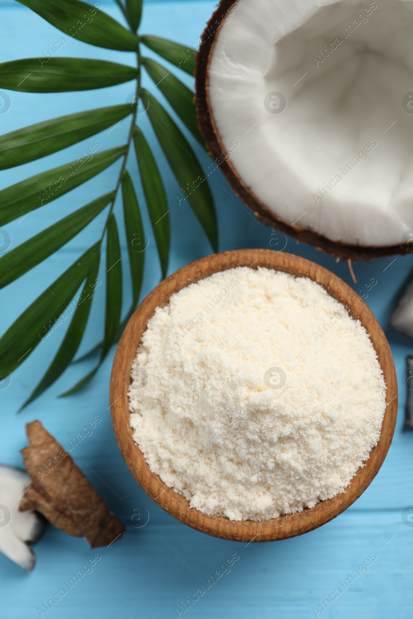 Photo of Fresh coconut flour in bowl, nut and palm leaf on light blue wooden table, flat lay