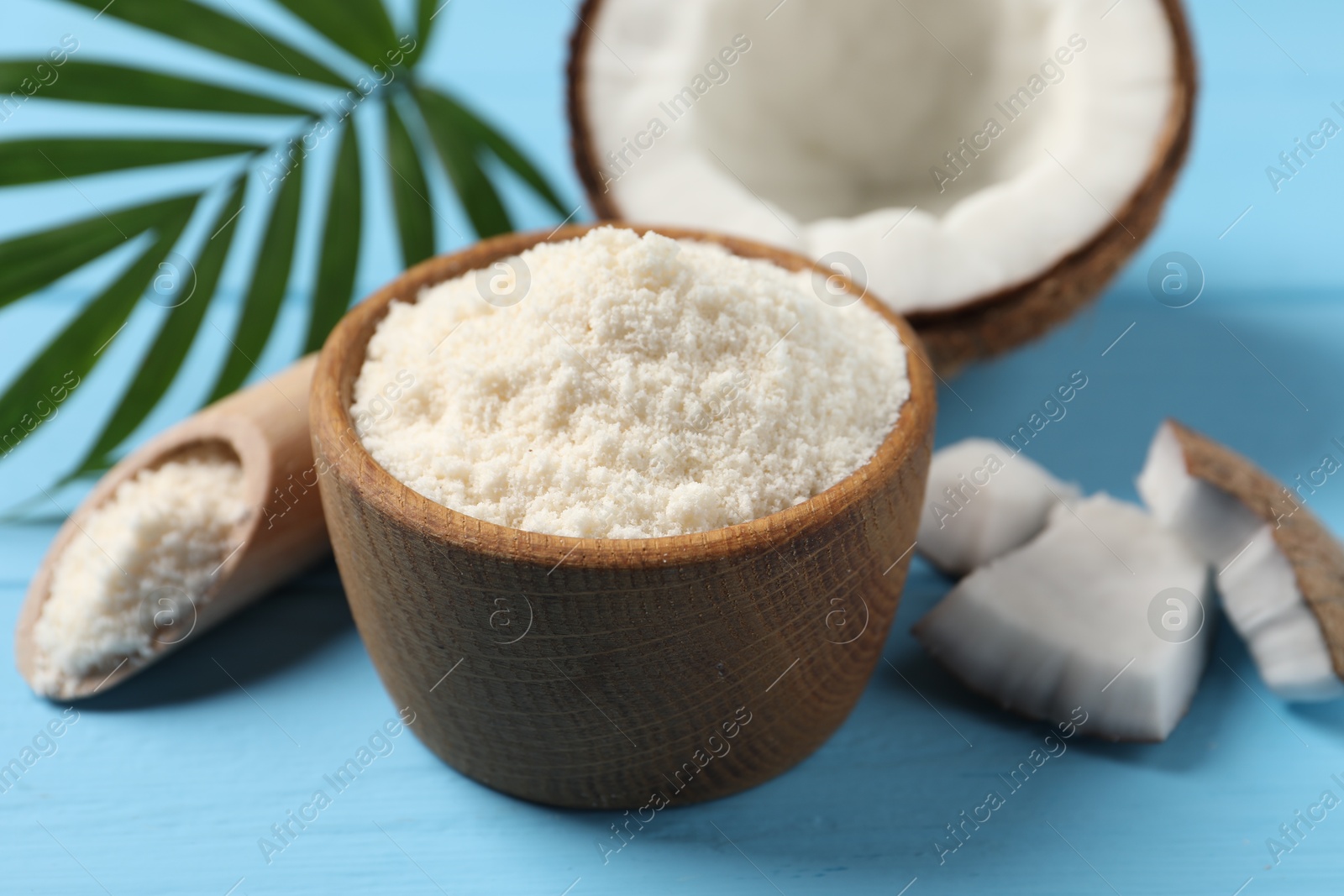 Photo of Fresh coconut flour in bowl, scoop, nut and palm leaf on light blue wooden table