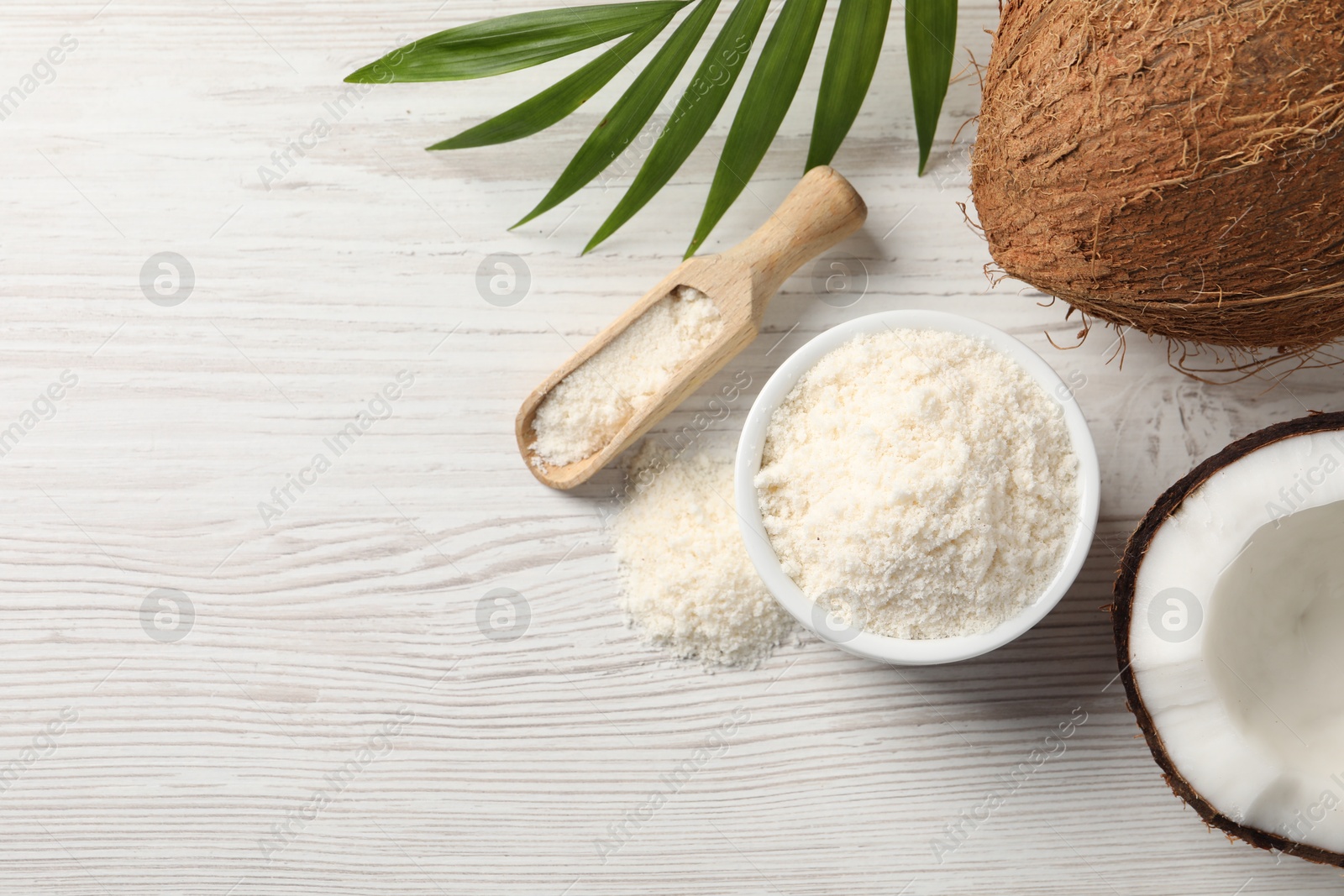 Photo of Fresh coconut flour in bowl, scoop, palm leaf and nuts on white wooden table, flat lay. Space for text