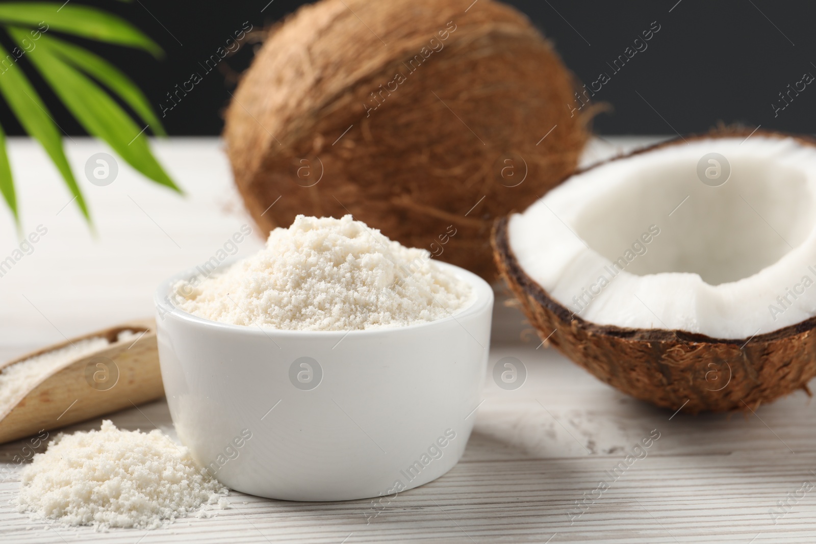 Photo of Fresh coconut flour in bowl, nuts, scoop and palm leaf on white wooden table