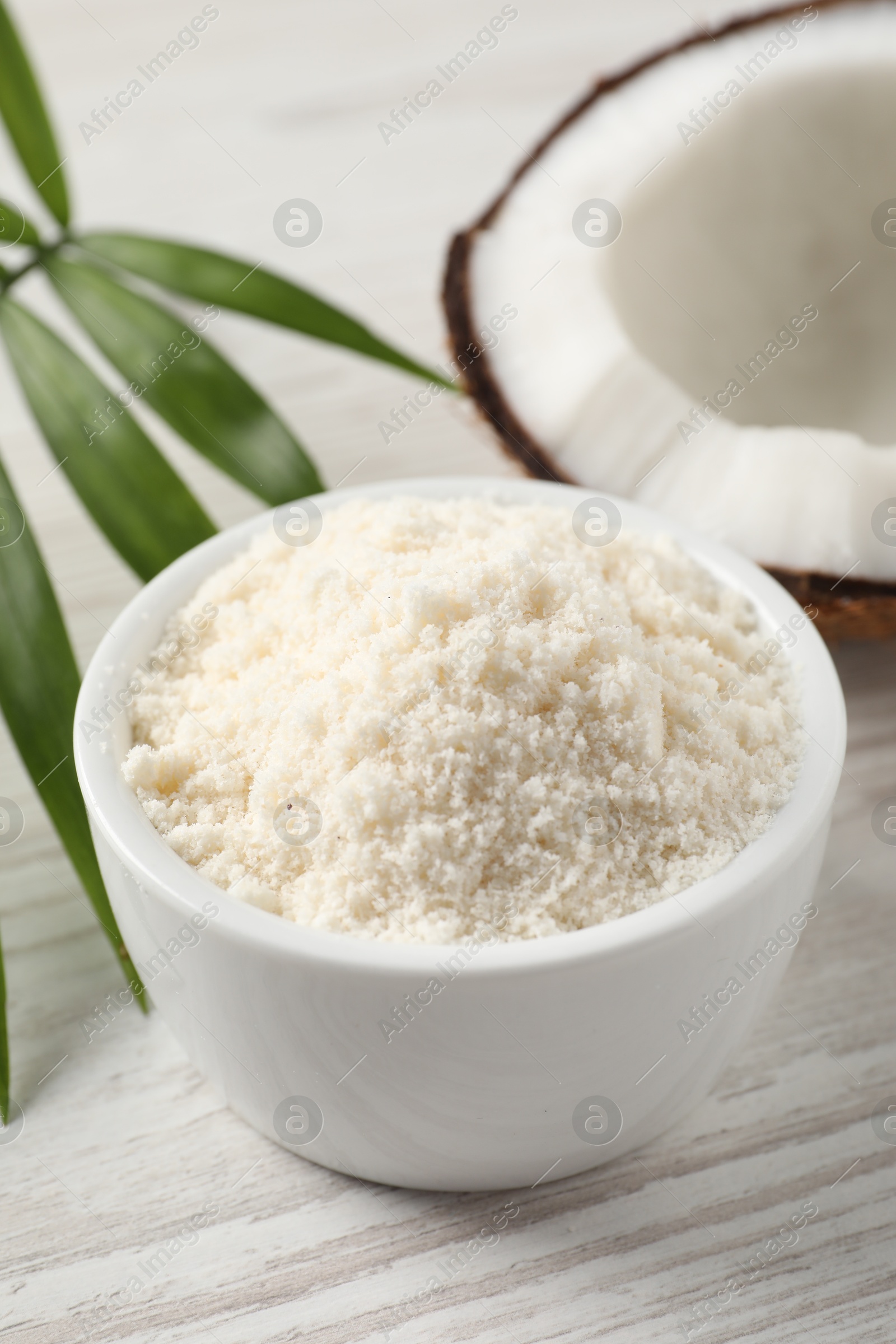 Photo of Fresh coconut flour in bowl, nut and palm leaf on white wooden table