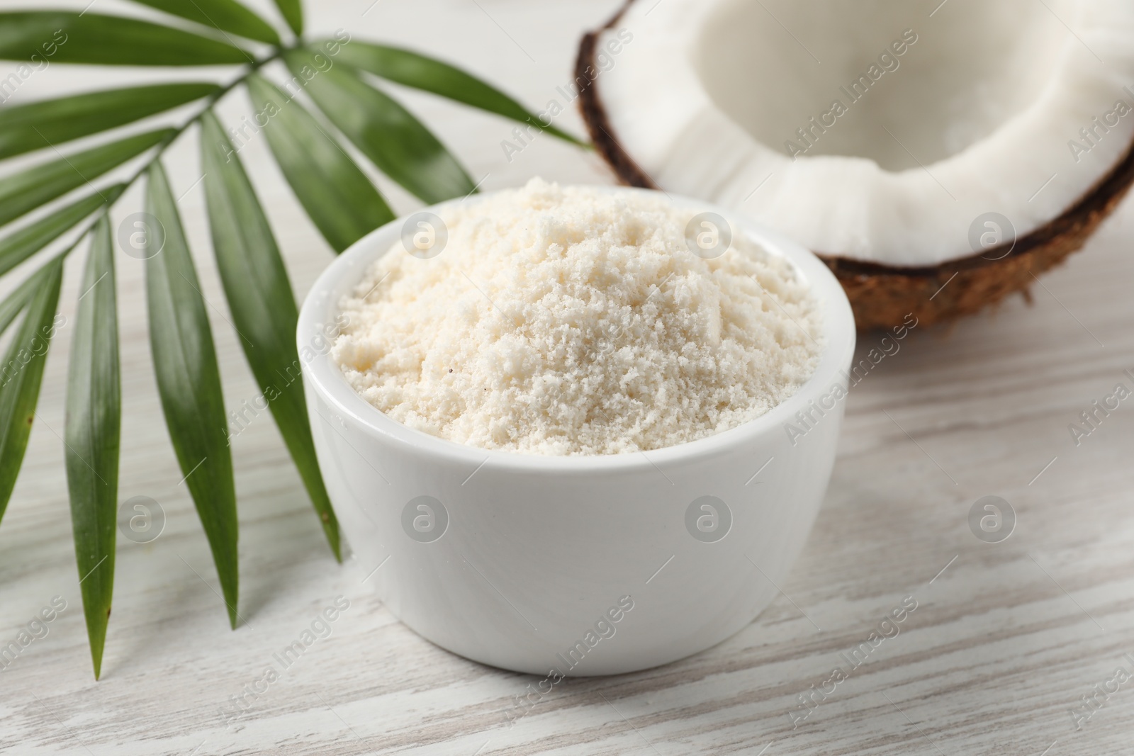 Photo of Fresh coconut flour in bowl, nut and palm leaf on white wooden table, closeup