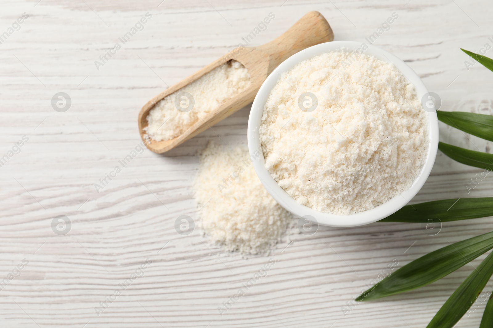 Photo of Fresh coconut flour in bowl, scoop and palm leaf on white wooden table, top view. Space for text