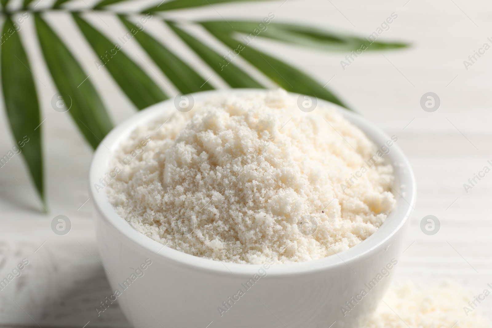 Photo of Fresh coconut flour in bowl and palm leaf on white table, closeup