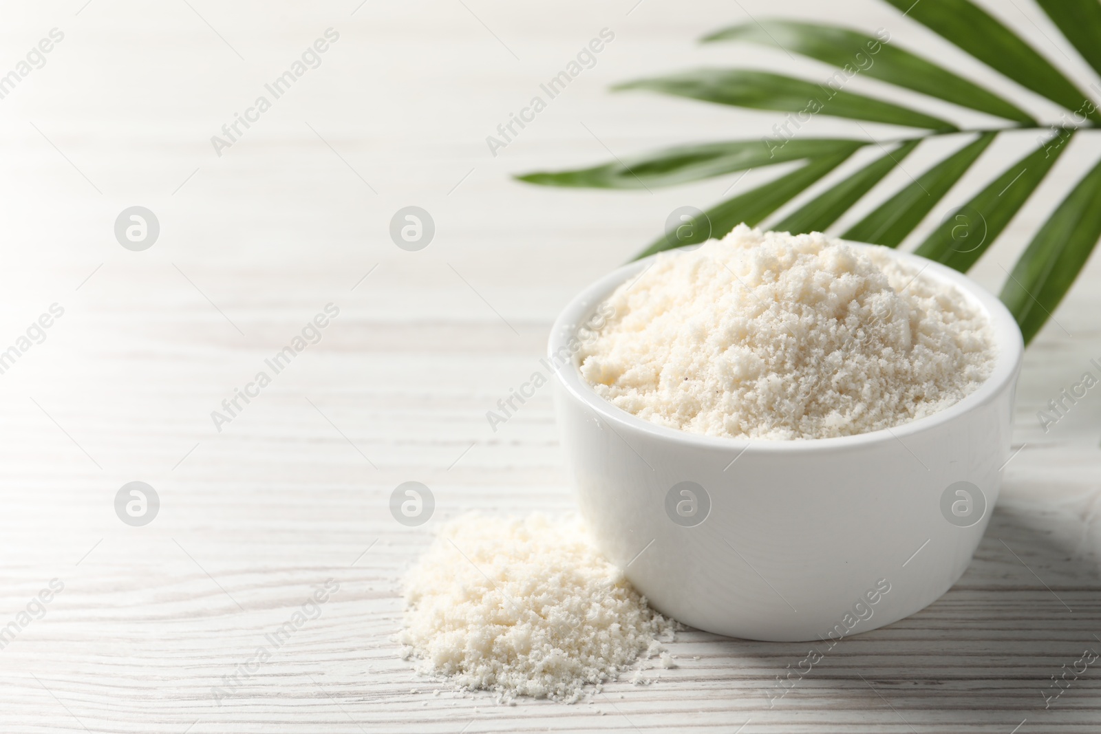 Photo of Fresh coconut flour in bowl and palm leaf on white wooden table, space for text