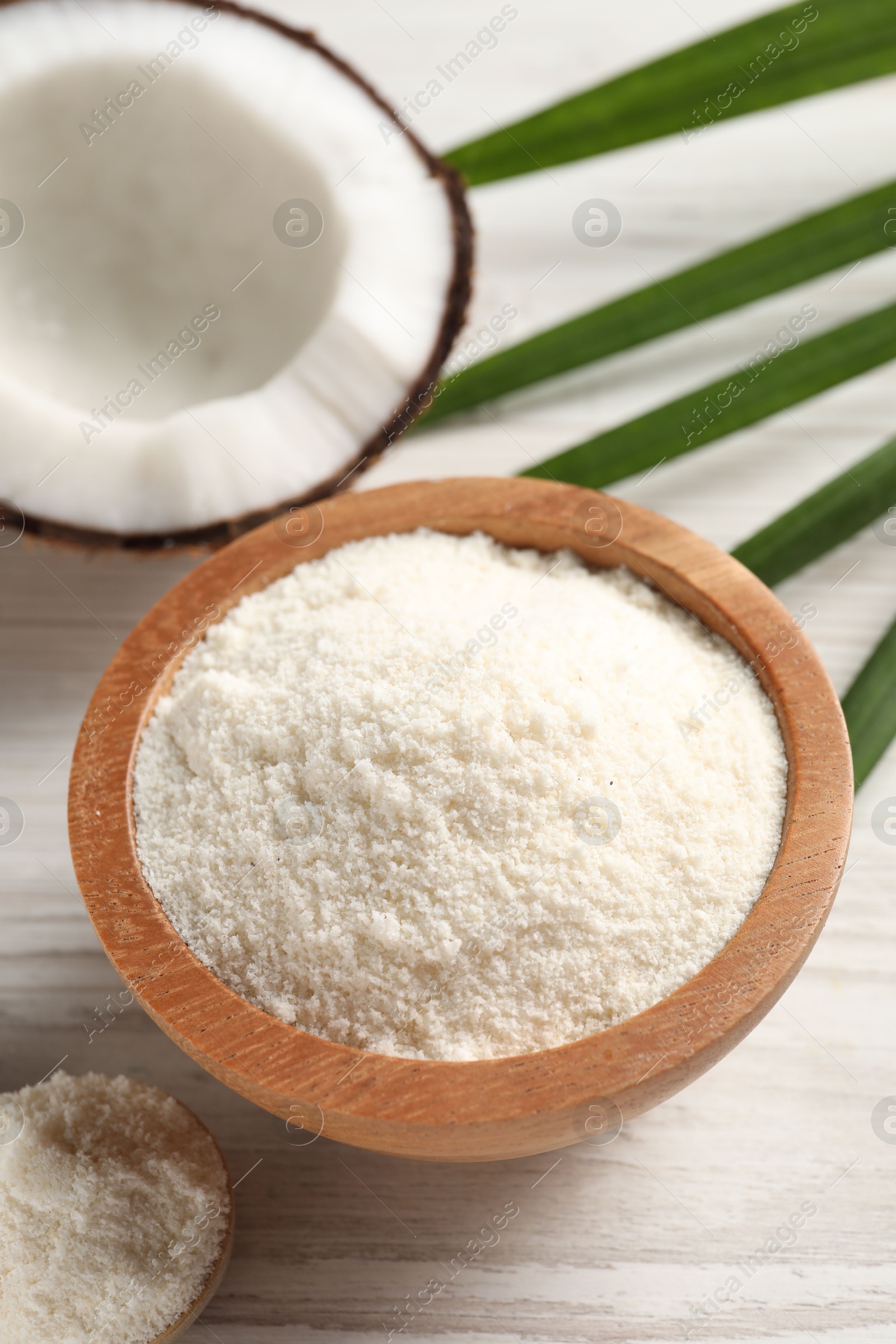 Photo of Fresh coconut flour in bowl, spoon, palm leaf and nut on white wooden table