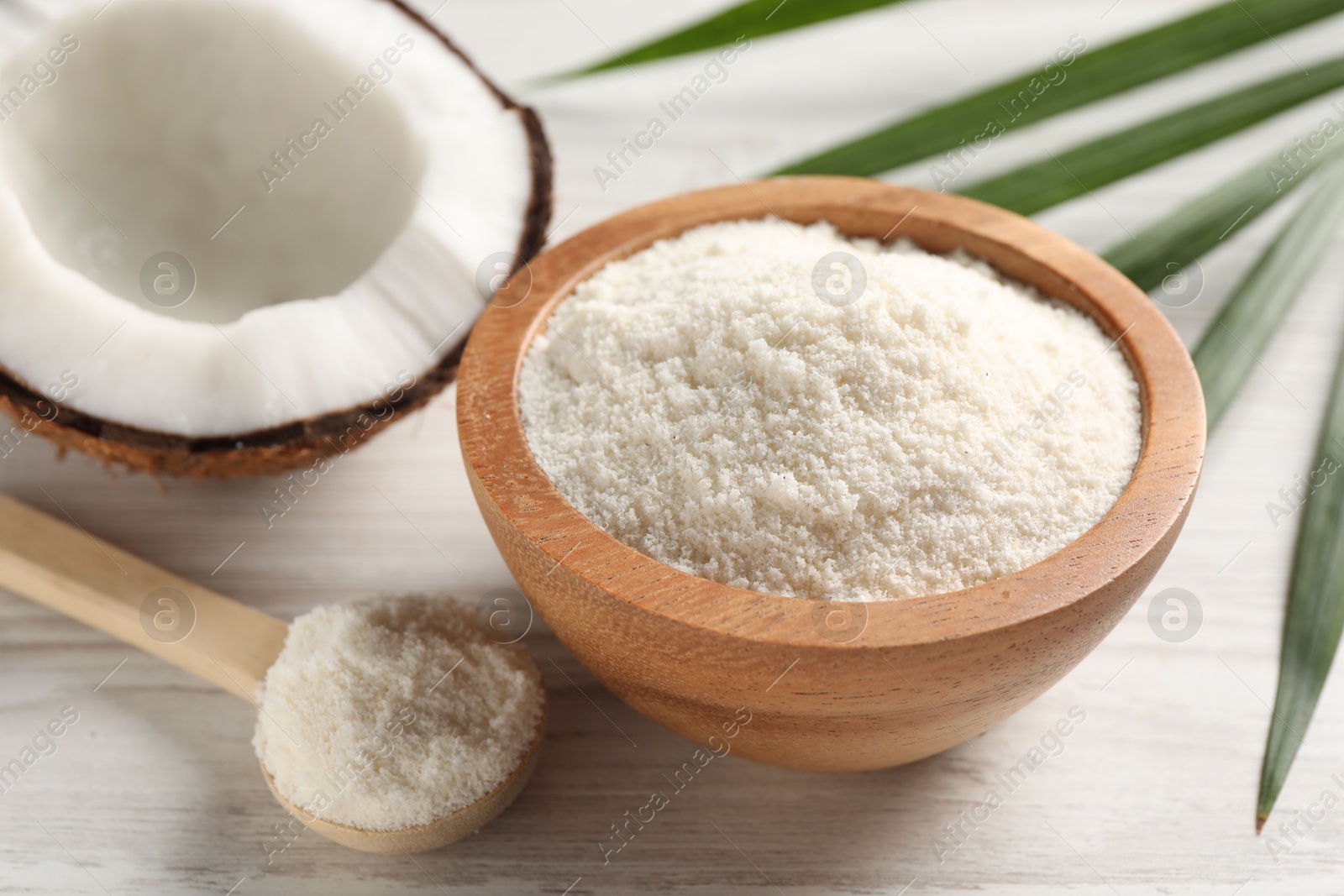 Photo of Fresh coconut flour in bowl, spoon, palm leaf and nut on white wooden table, closeup