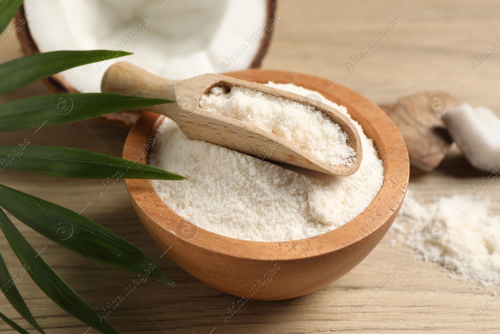 Photo of Fresh coconut flour in bowl, scoop, nut and palm leaf on wooden table, closeup