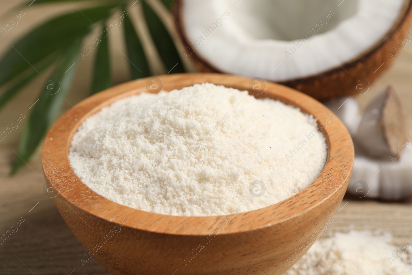 Photo of Fresh coconut flour in bowl, nut and palm leaf on white wooden table, closeup