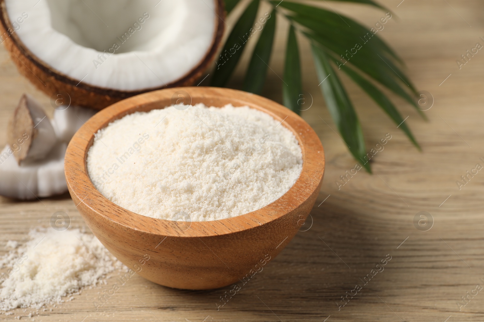 Photo of Fresh coconut flour in bowl, nut and palm leaf on white wooden table, closeup