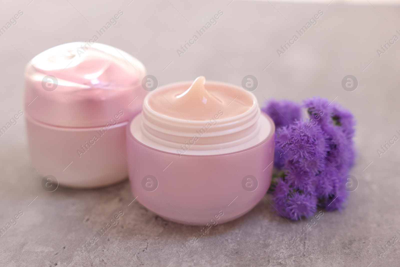 Photo of Jars of creams and ageratum flowers on gray table, closeup