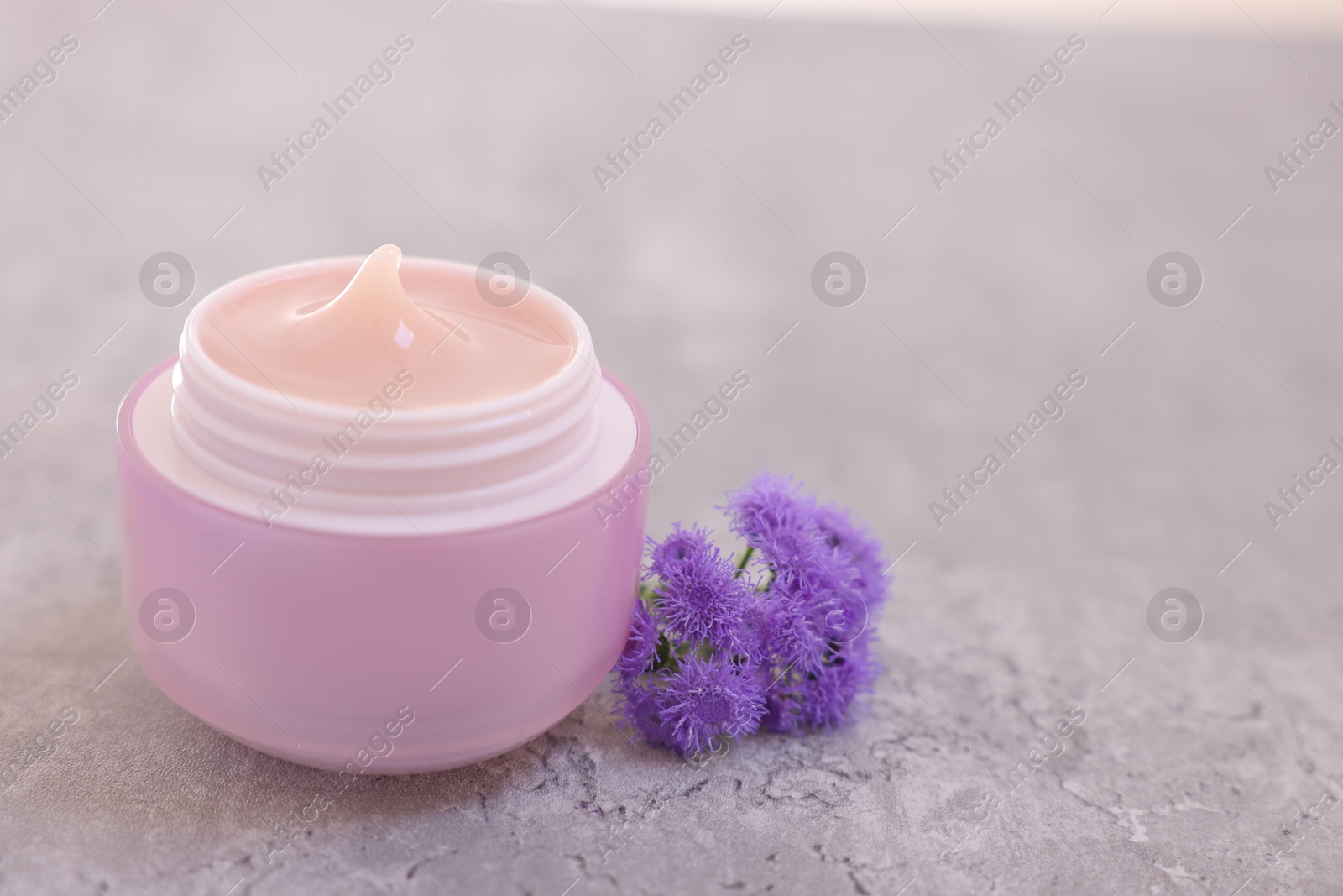 Photo of Jar of cream and ageratum flowers on gray table, closeup. Space for text