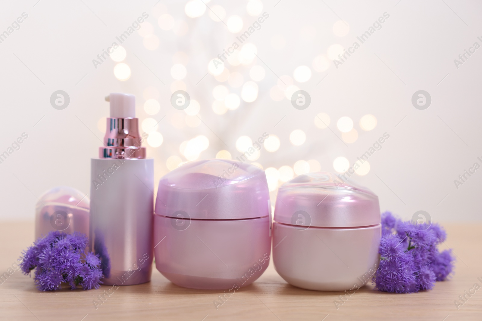 Photo of Set of different cosmetic products and ageratum flowers on wooden table against light background with blurred lights