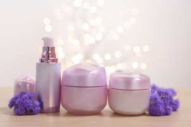 Photo of Set of different cosmetic products and ageratum flowers on wooden table against light background with blurred lights