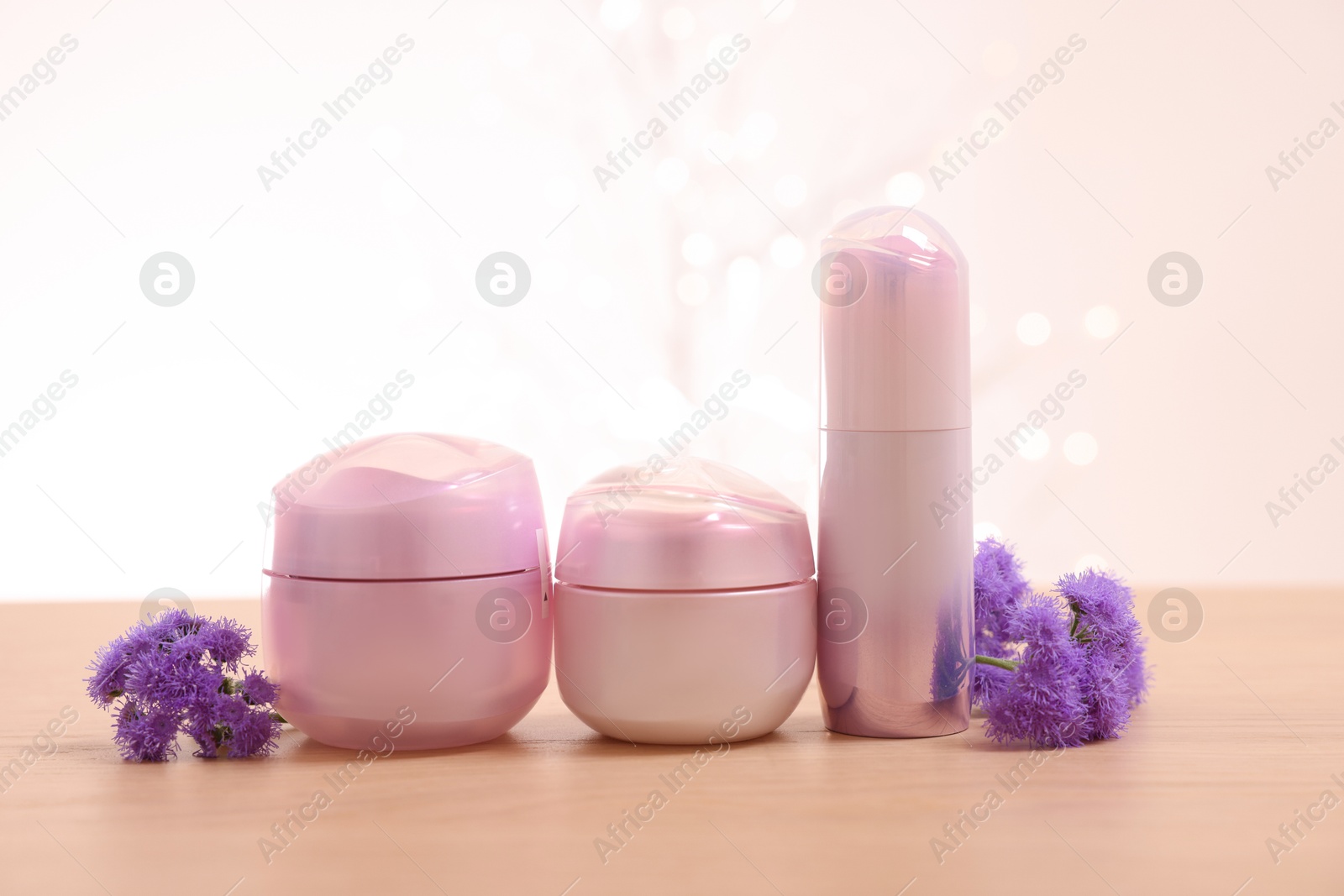 Photo of Set of different cosmetic products and ageratum flowers on wooden table against light background with blurred lights