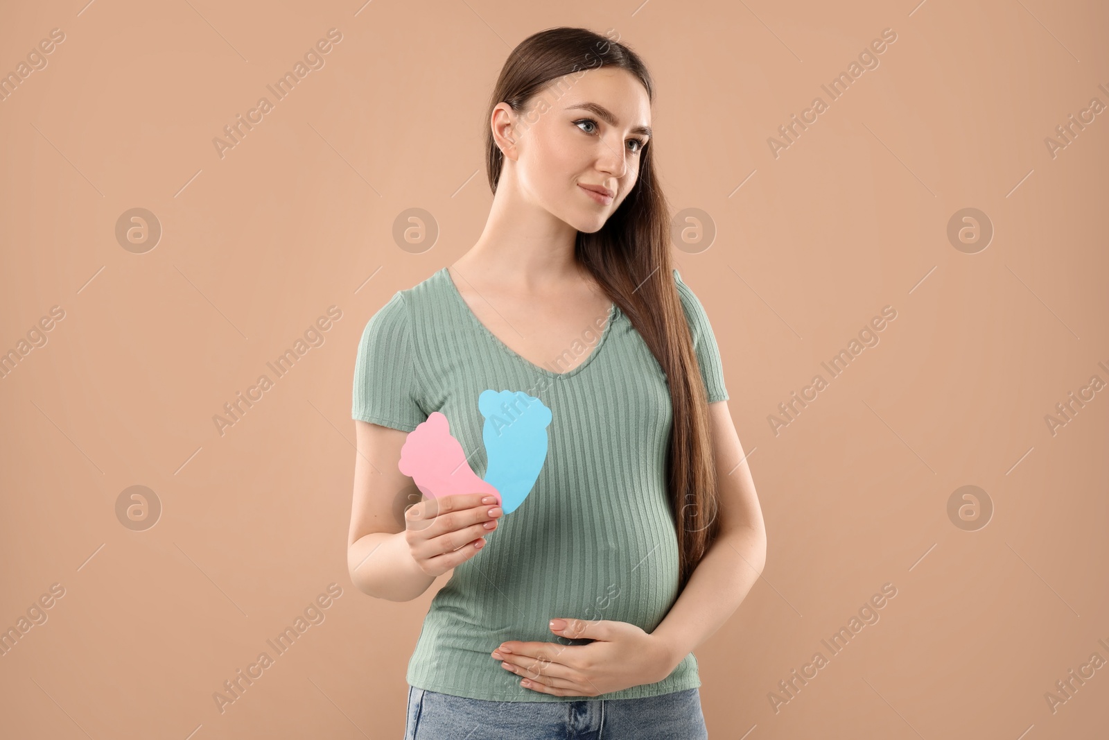 Photo of Expecting twins. Pregnant woman holding two paper cutouts of feet on light brown background