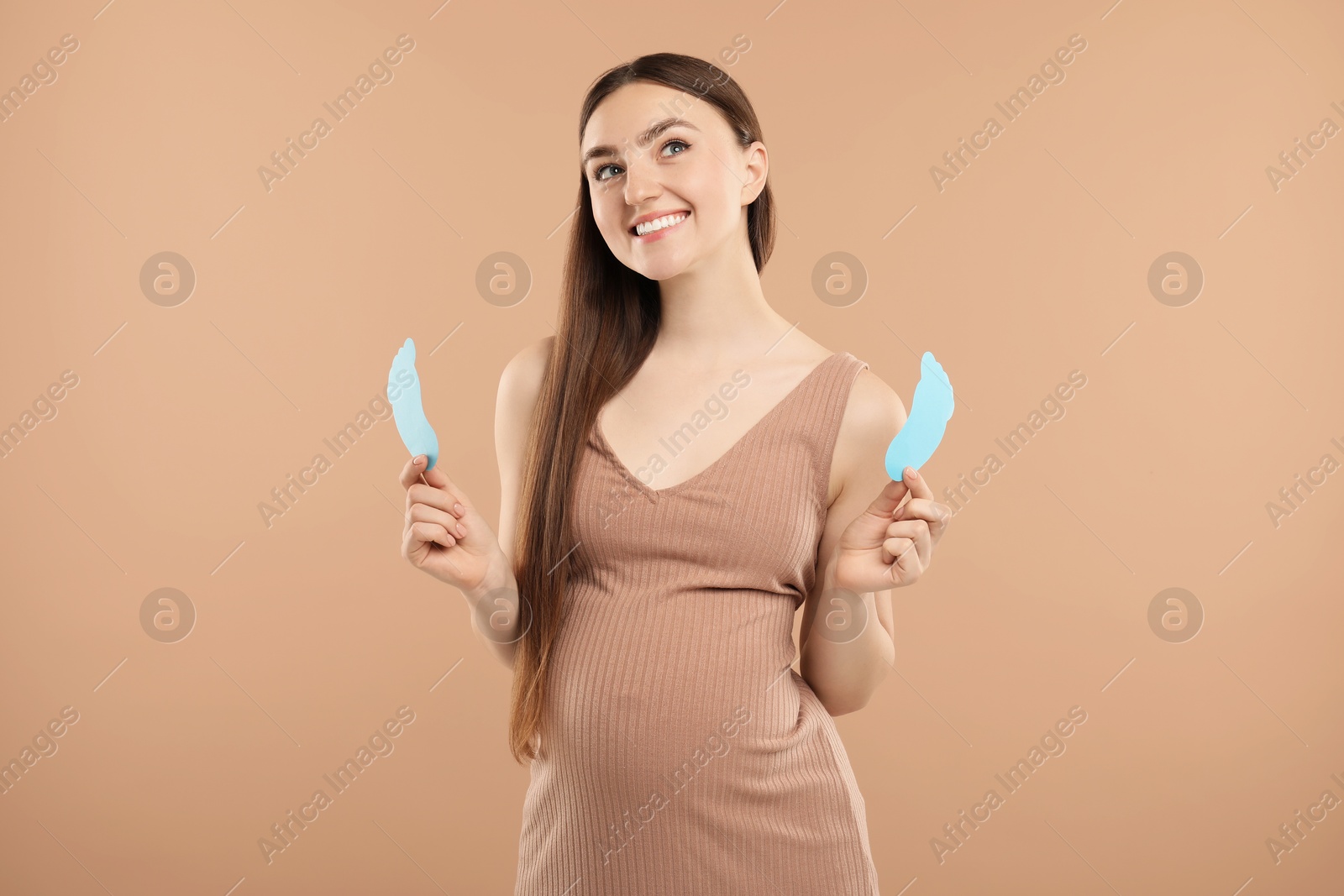 Photo of Expecting twins. Pregnant woman holding two paper cutouts of feet on light brown background