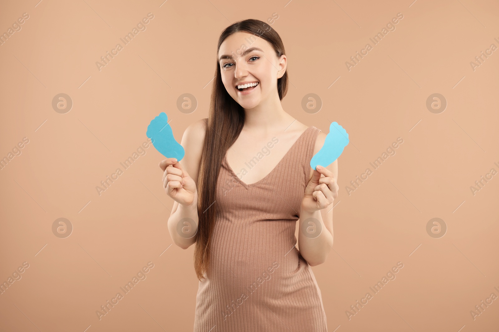 Photo of Expecting twins. Pregnant woman holding two paper cutouts of feet on light brown background