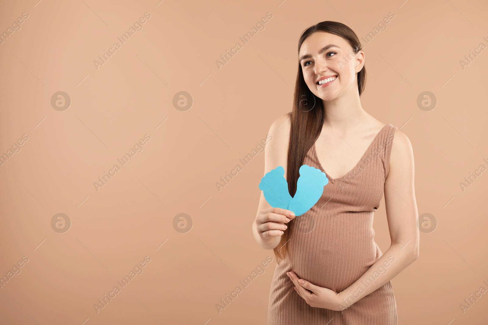 Photo of Expecting twins. Pregnant woman holding two paper cutouts of feet on light brown background, space for text