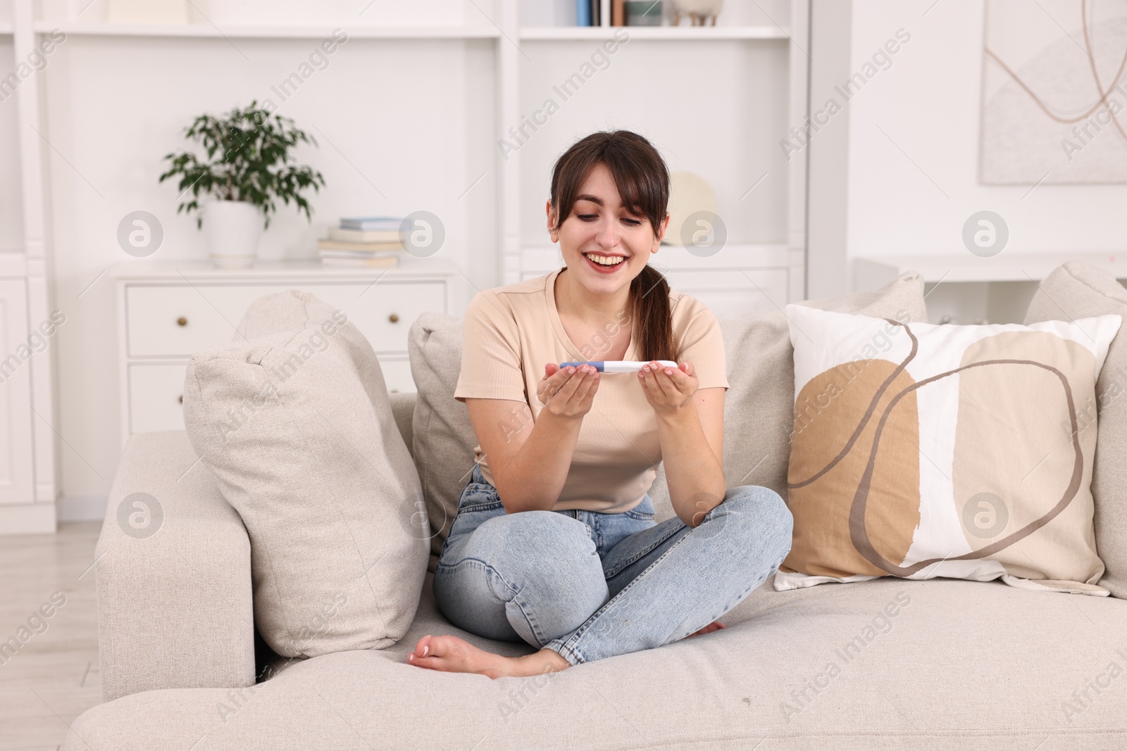 Photo of Happy young woman with pregnancy test on sofa at home