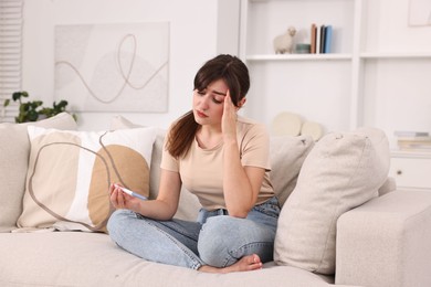 Photo of Worried young woman with pregnancy test on sofa at home