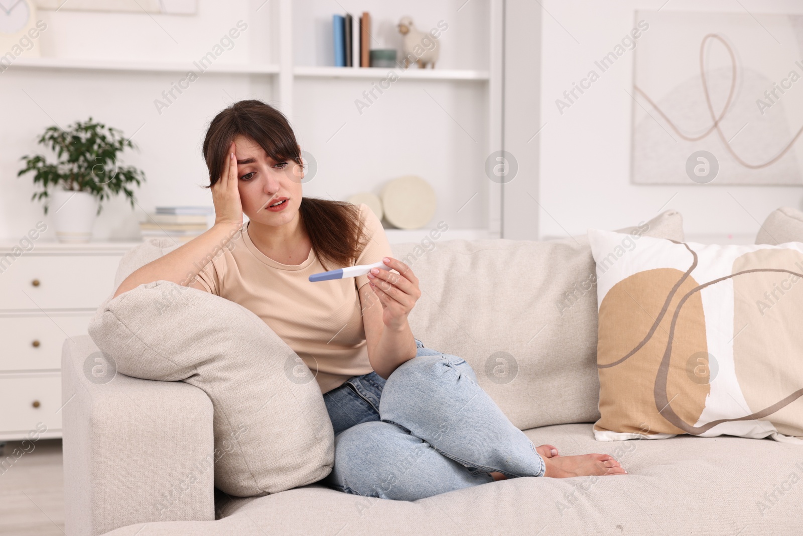 Photo of Worried young woman with pregnancy test on sofa at home