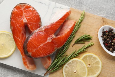 Board with fresh salmon steaks, peppercorns, rosemary and lemon on grey textured table, top view