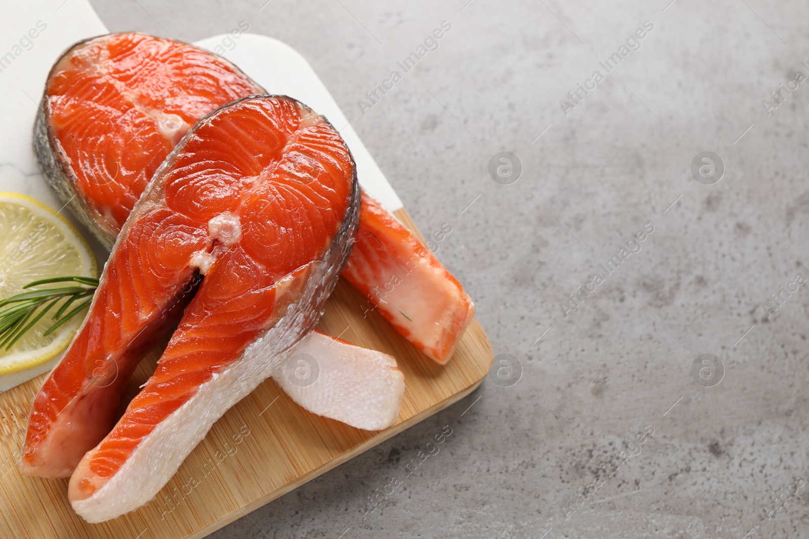 Photo of Board with fresh salmon steaks, rosemary and lemon on grey textured table, closeup. Space for text