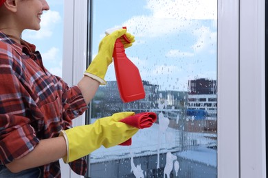 Photo of Woman with spray bottle of detergent and napkin cleaning window indoors, closeup