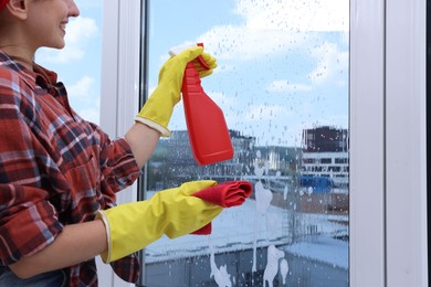 Woman with spray bottle of detergent and napkin cleaning window indoors, closeup