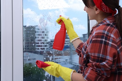 Photo of Woman with spray bottle of detergent and napkin cleaning window indoors, closeup