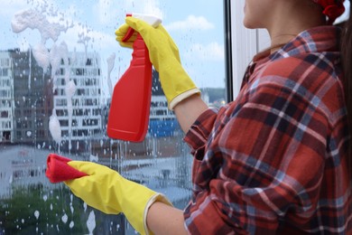 Woman with spray bottle of detergent and napkin cleaning window indoors, closeup